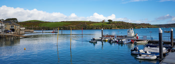 Salcombe harbour This is a landscape photograph capturing Salcombe harbour in Devon, England during the early afternoon in winter. The image prominently features the harbour's tranquil waters, with several boats docked along floating pontoons, highlighting transport activity typical of this coastal area. Traditional stone buildings are visible along the waterfront, set against green hills that rise beyond the harbour, illustrating the town's scenic locale in the United Kingdom. The photograph provides a clear view of the boats that serve both leisure and practical transport purposes in Salcombe, exemplifying the connection between local life and maritime activities in this part of England.